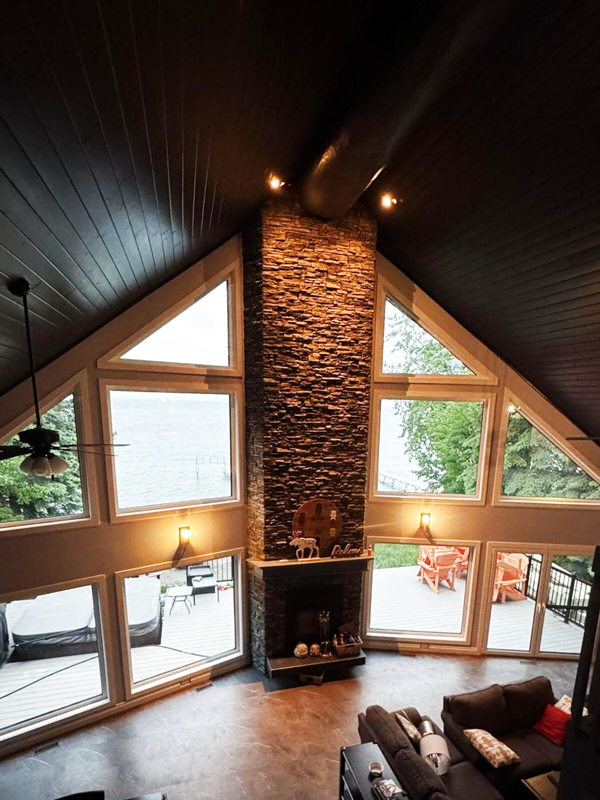 A-frame great room at dusk — stone chimney flanked by gable and picture windows looking onto the lake